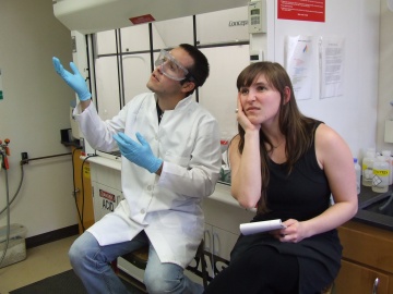 A young white man wearing a lab coat, gloves and goggles is busily explaining science to a young white female with a notepad who looks bored and discouraged. The conversation is taking place in a lab.